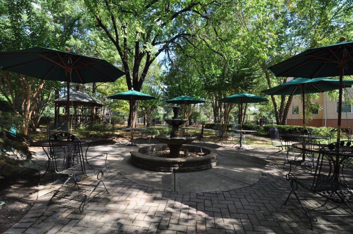 A serene outdoor patio with a central fountain, surrounded by tables, chairs, and green umbrellas under the shade of trees.