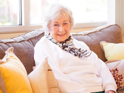An elderly woman sitting comfortably on a sofa, smiling warmly.