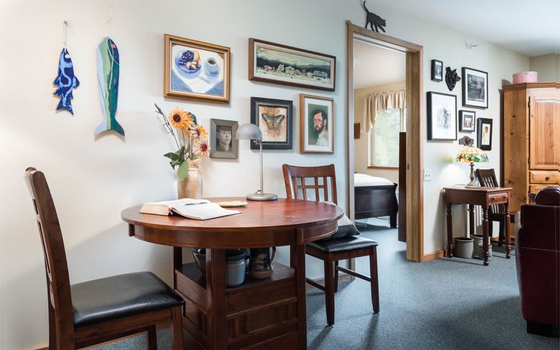 A cozy interior space featuring a round dining table with chairs, surrounded by various artworks and decorations on the wall. A vase of sunflowers adds a touch of freshness to the room.