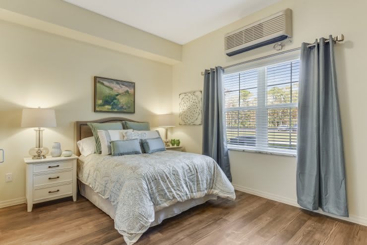 A cozy bedroom with a neatly made bed, curtains drawn back from a window and an air conditioner above.