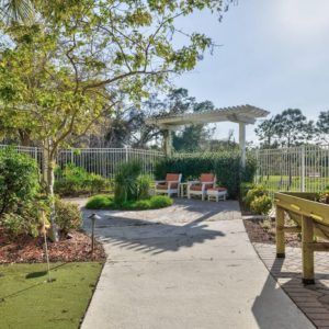 A serene garden pathway leads to a shaded seating area with chairs, surrounded by lush greenery on a sunny day.
