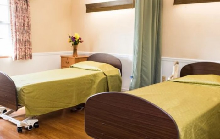 A tidy hospital room featuring two beds with green covers, a bedside table with flowers, and privacy curtains.