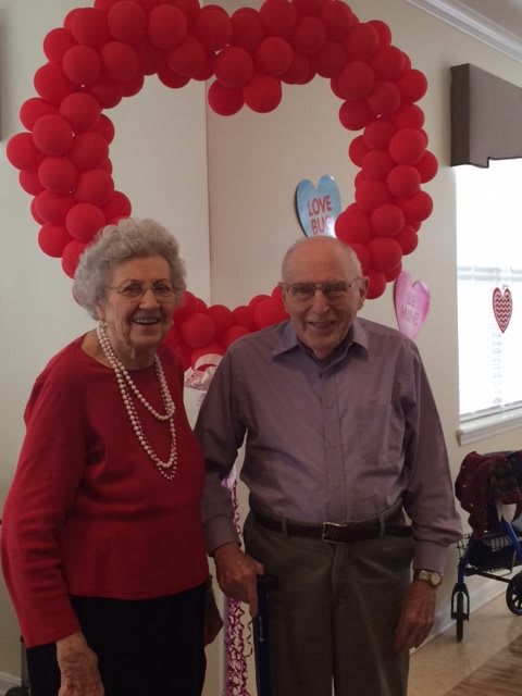 An elderly couple standing together in front of heart-shaped balloon decorations.