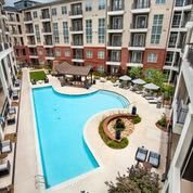 A view of an apartment complex with a spacious outdoor swimming pool surrounded by landscaped greenery and patio furniture.