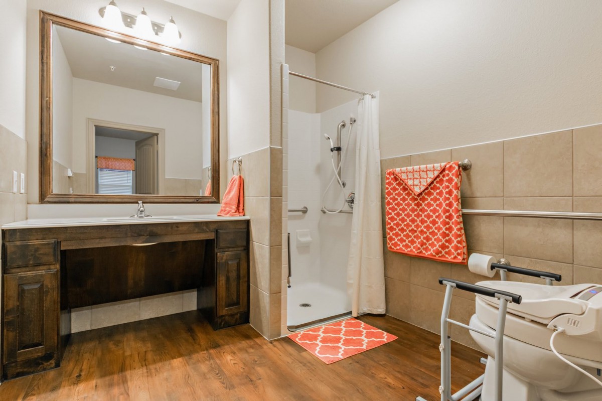A modern accessible bathroom with a walk-in shower, wooden flooring, and orange towels.