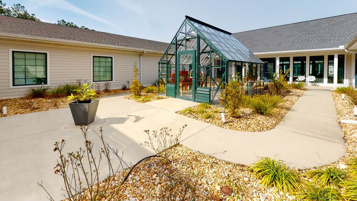 A small greenhouse in a garden setting, surrounded by various plants and pathways.