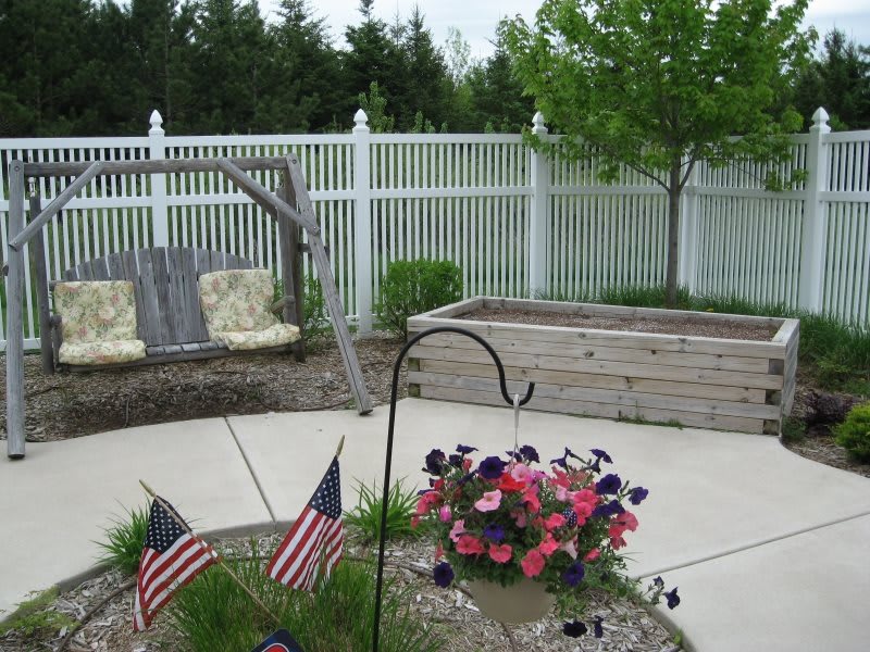 A peaceful garden patio with a swing, a flower pot adorned with small flags, and a white fence in the background.