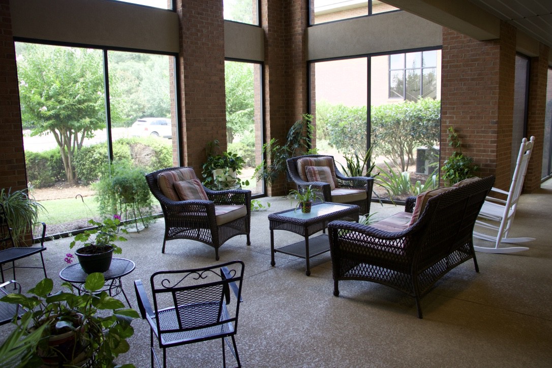 A cozy lounge area featuring wicker furniture, surrounded by large windows and lush indoor plants.