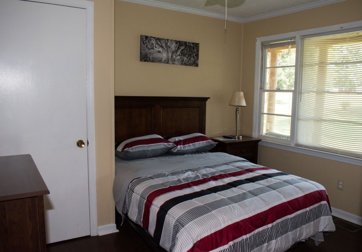 A cozy bedroom featuring a neatly made bed with striped bedding, a nightstand with a lamp, and a window allowing natural light to filter in.