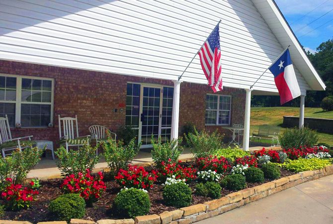 A house with a neatly landscaped garden featuring colorful flowers, patio chairs, and two flags displayed.