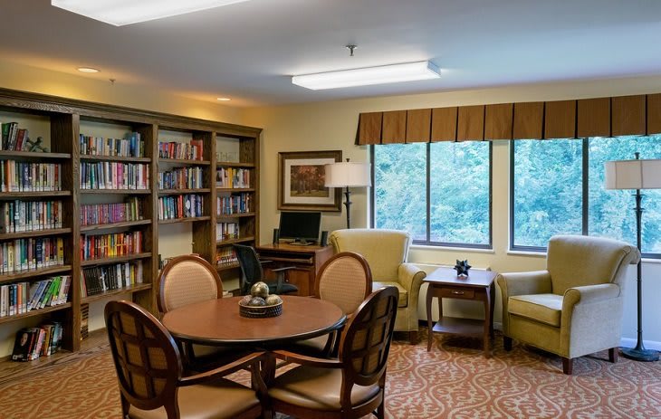 A cozy library space with a round table, chairs, bookshelves, and large windows.