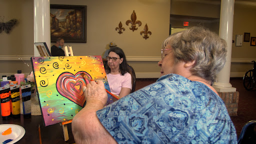 An elderly woman focuses on painting a colorful heart on her canvas while others engage in a creative art session.