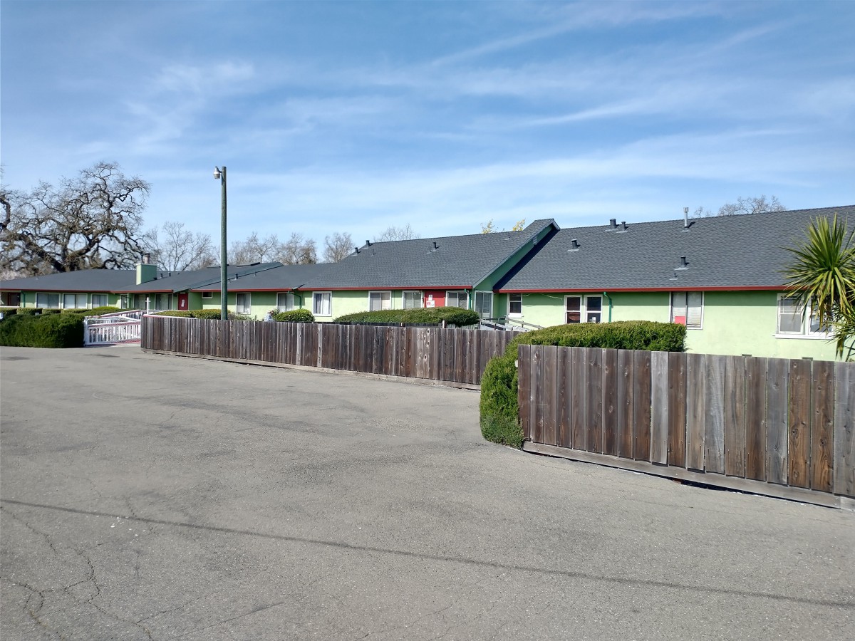 A row of motel buildings with a wooden fence alongside a wide road under a clear blue sky.