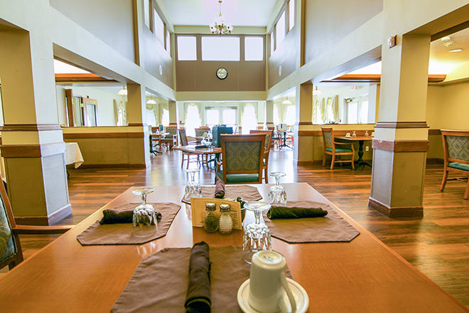 A spacious dining hall with neatly arranged tables and chairs, featuring a wooden floor and large windows.