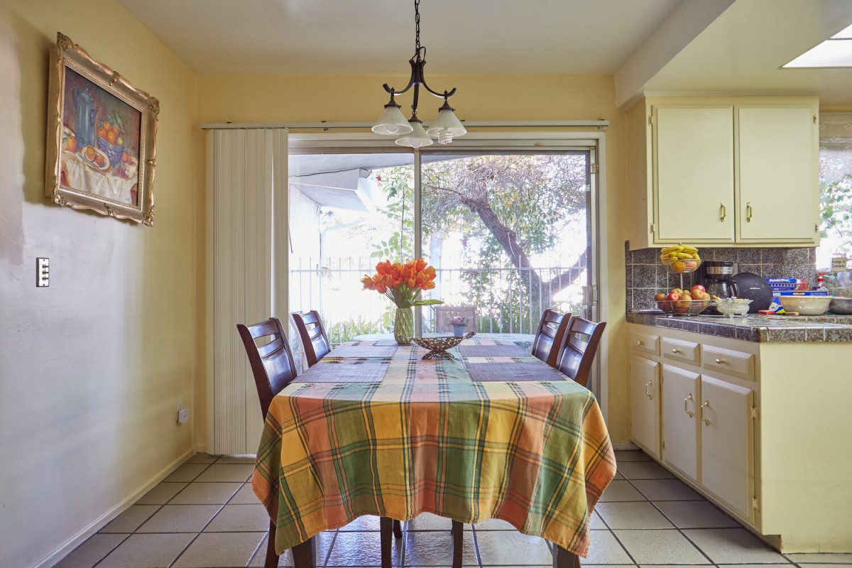 A cozy dining area with a colorful tablecloth and flowers on the table, well-lit by natural sunlight through large windows.