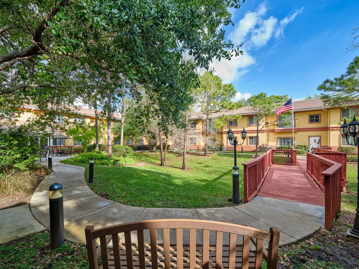 A tranquil garden with a wooden bench, surrounded by trees and buildings under a clear blue sky.