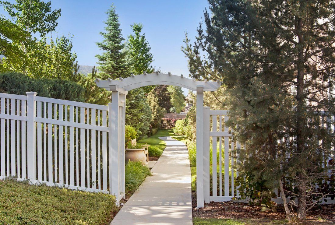 A scenic garden pathway with a white arch and fence, surrounded by lush greenery.