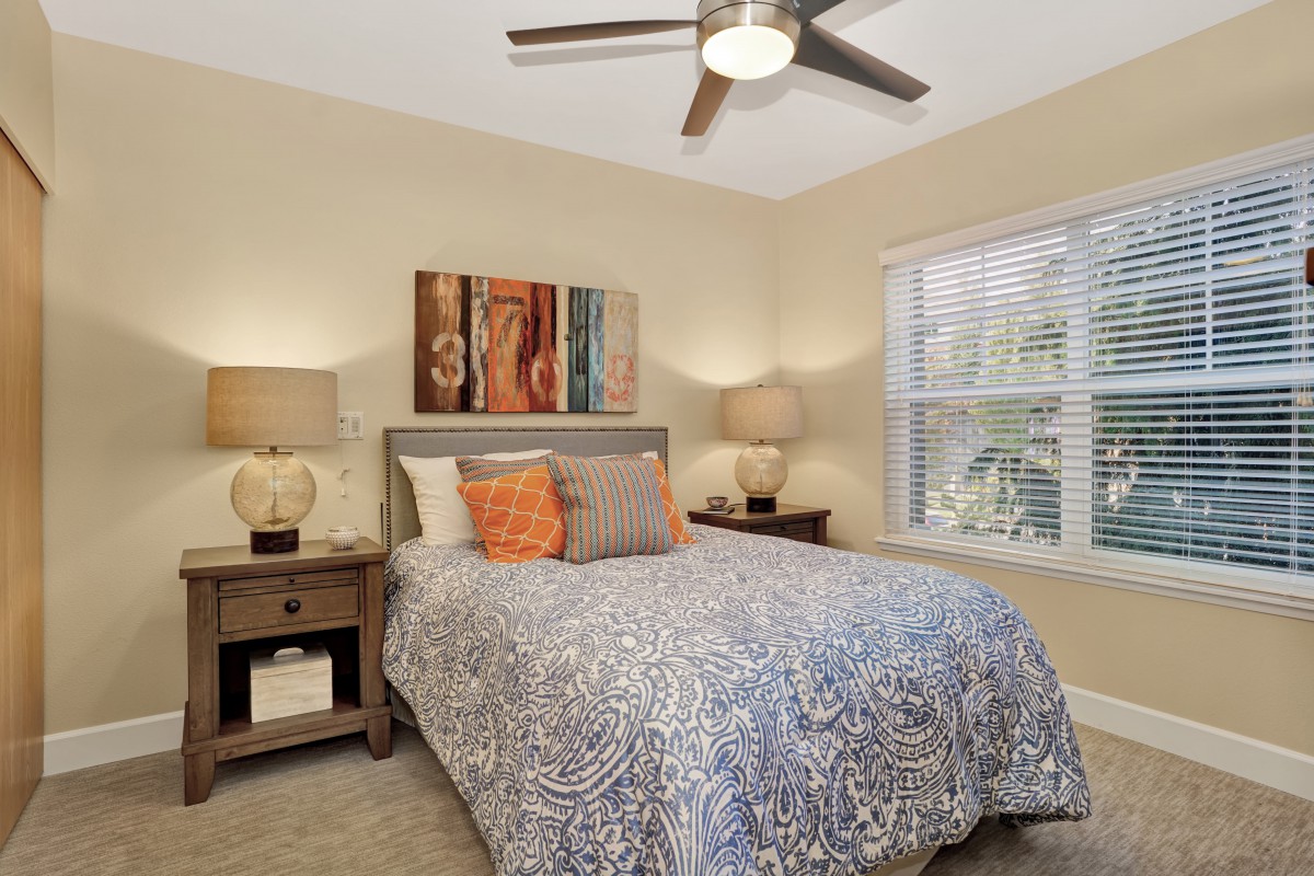 A cozy bedroom features a neatly arranged bed with patterned bedding, flanked by two lamps on nightstands. Artwork adorns the wall above the headboard, and natural light filters through a window with blinds.