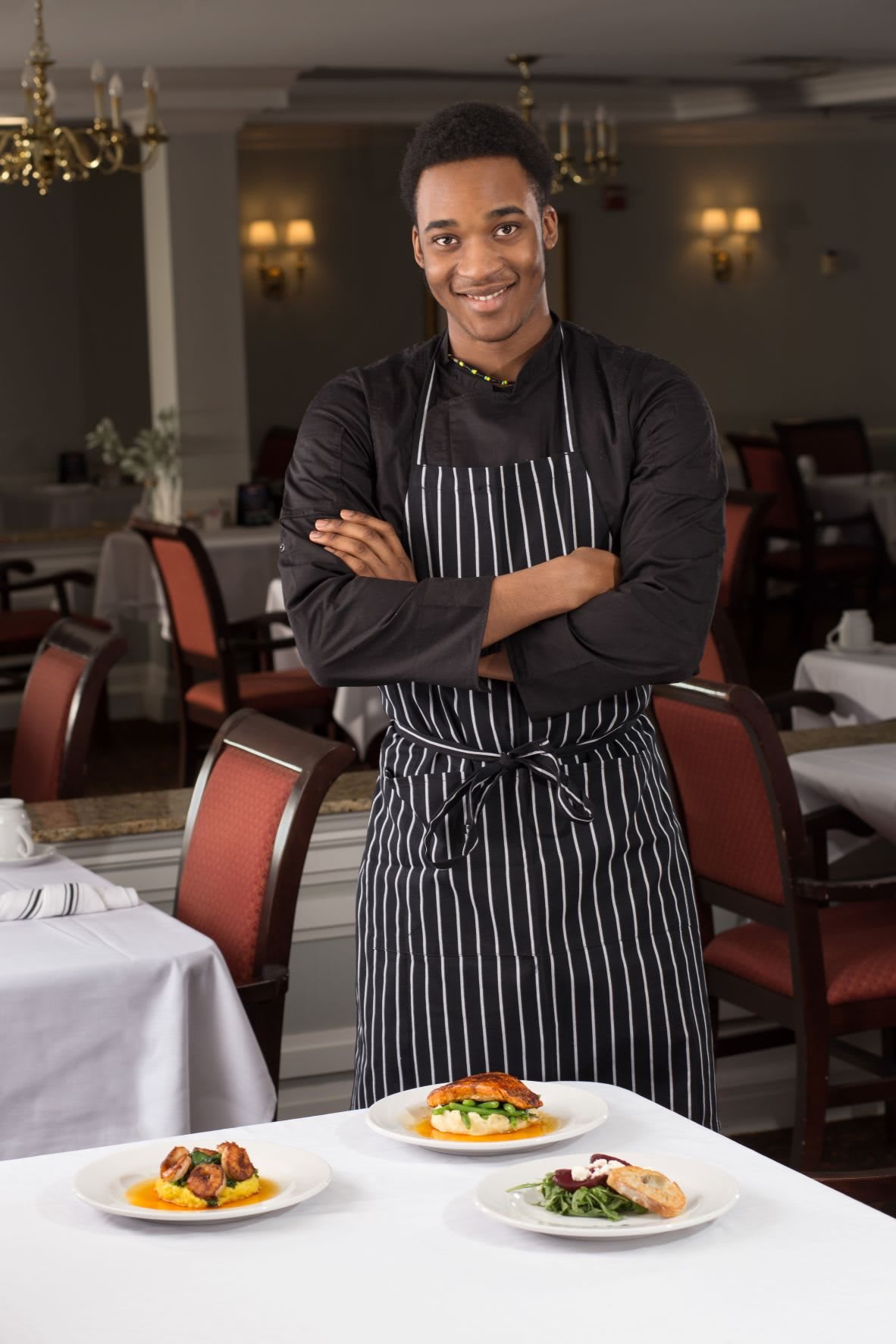 A smiling chef in an apron stands in a restaurant dining area with a prepared dish on the table.