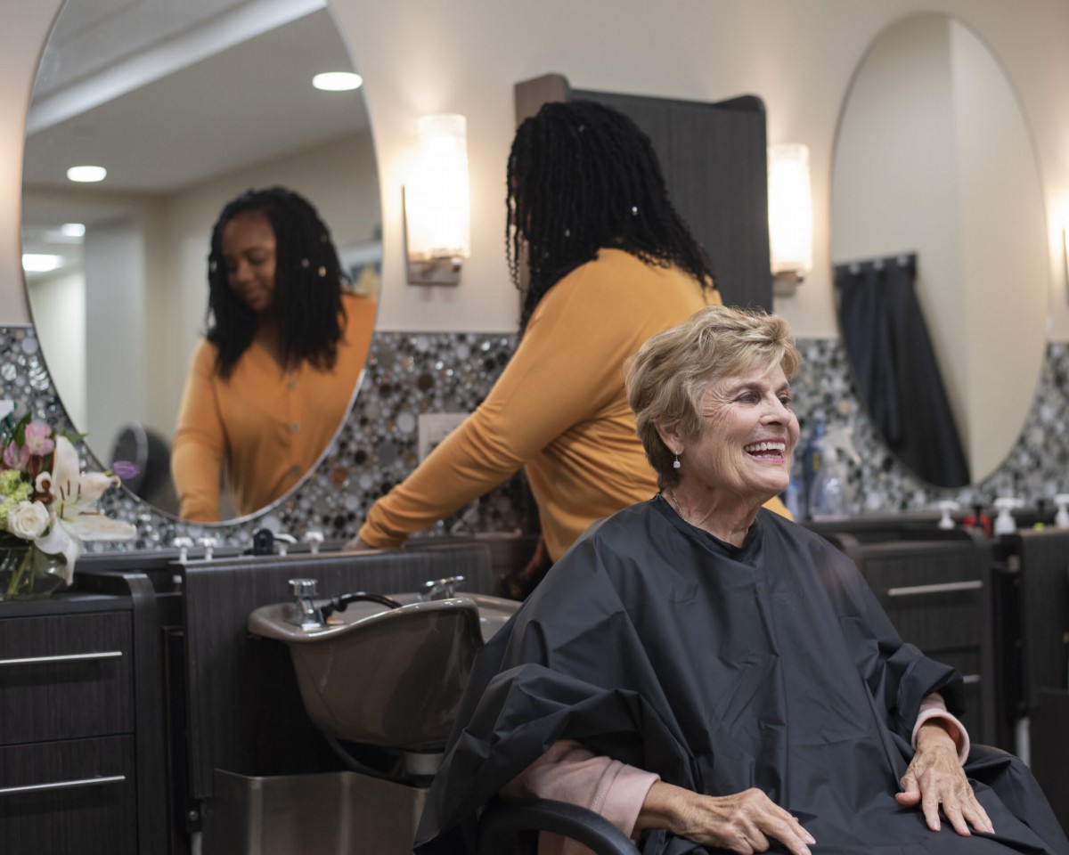 An elderly woman sits smiling in a hair salon chair while a hairdresser prepares behind her.
