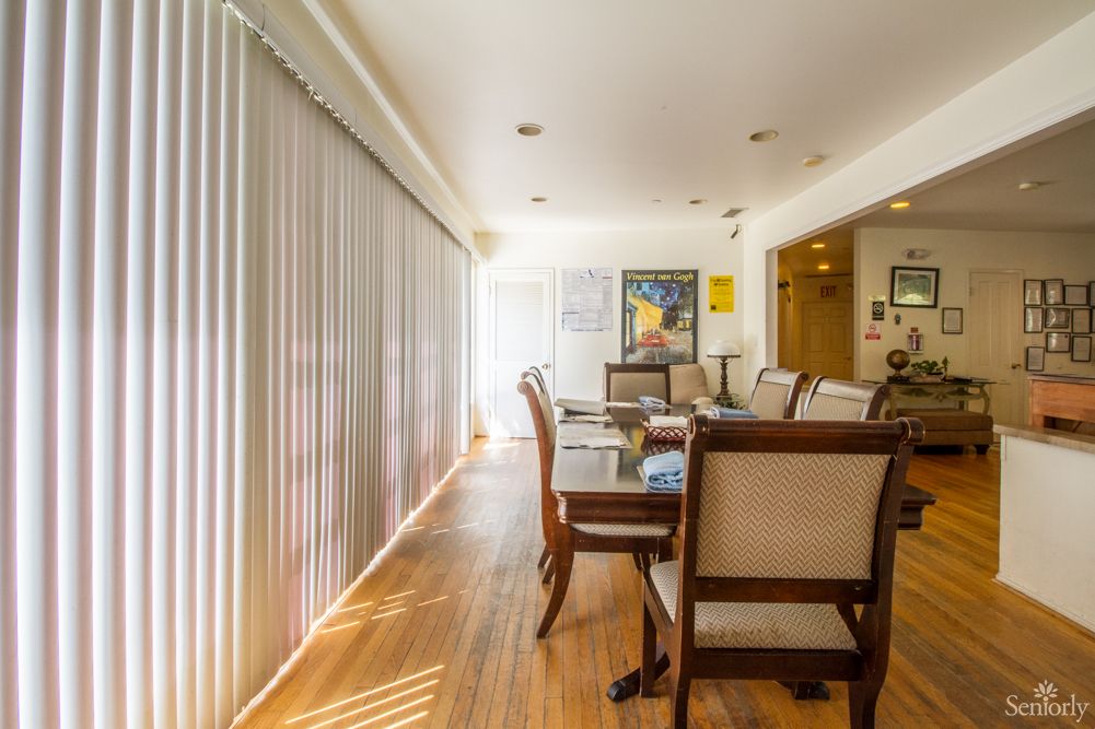 A well-lit dining room with a glass table and upholstered chairs, featuring natural light streaming through vertical blinds.