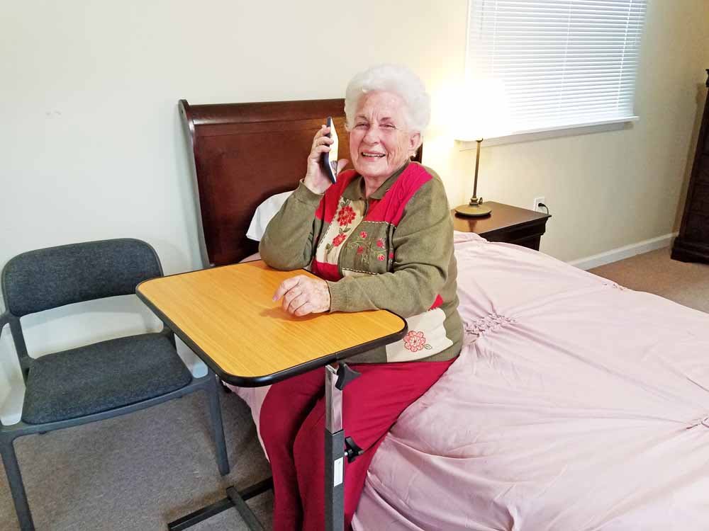 A smiling senior woman sits by a bed, talking on the phone with a mobile table in front of her.