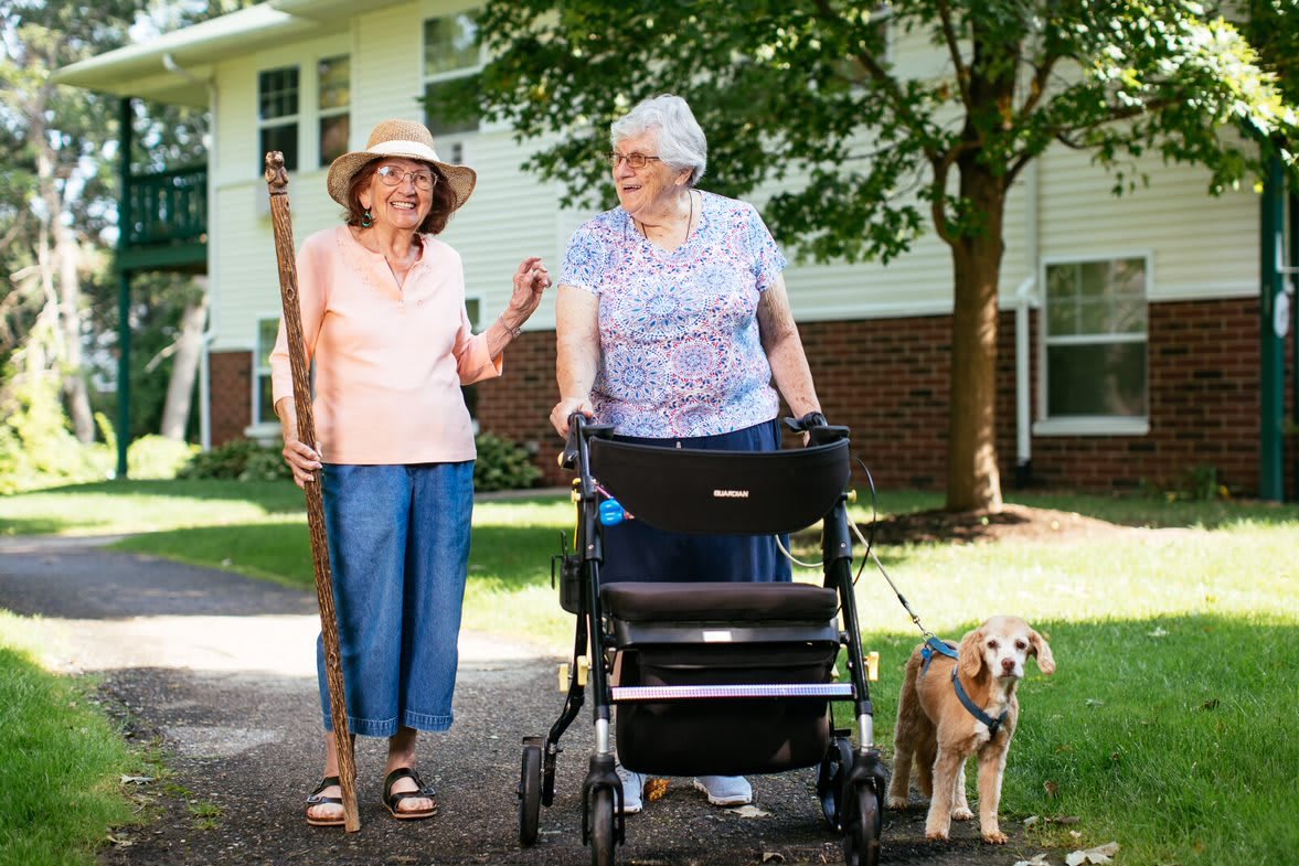 Two elderly women enjoying a walk outdoors with a dog, one using a cane and the other a walker.