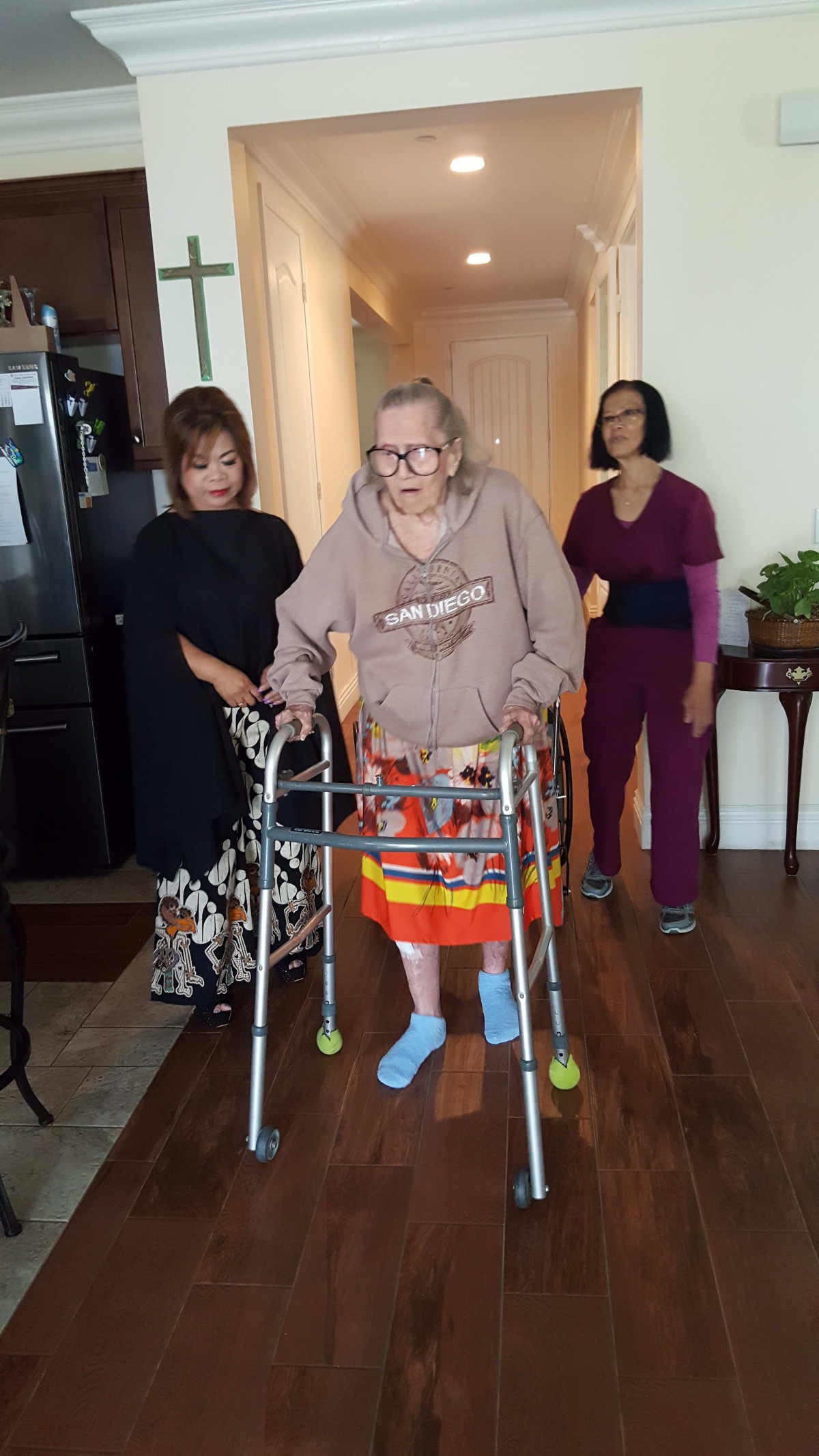 An elderly person using a walker, assisted by two caregivers in an indoor setting.