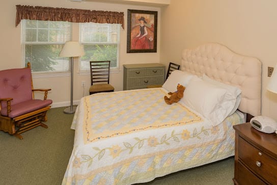 A cozy bedroom with a neatly made bed featuring a teddy bear, a lamp, and simple decor.