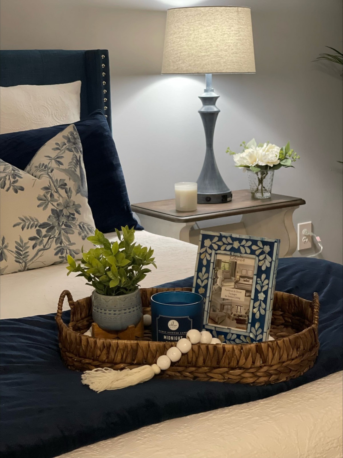 A cozy bedroom scene featuring a stylish tray with decor items, including a plant and picture frame, beside a softly lit lamp on the nightstand.