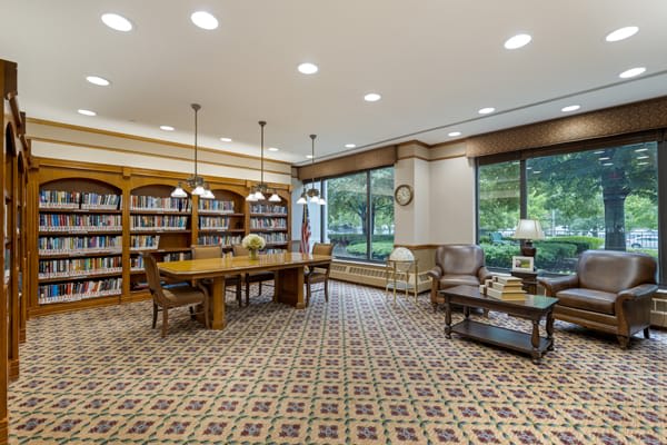 A cozy library reading room with bookshelves, a central wooden table, and large windows.