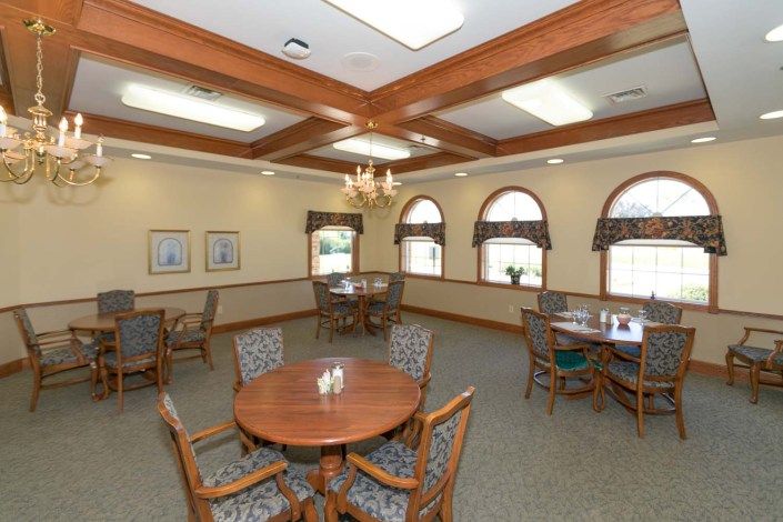 A well-lit dining room featuring round wooden tables, cushioned chairs, large windows, and a central chandelier.