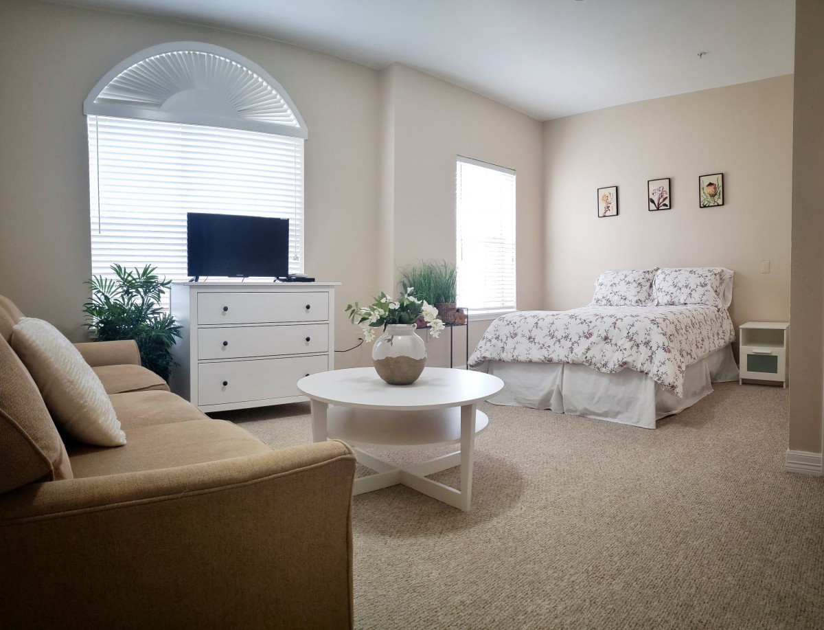 A cozy bedroom with a bed, sofa, dresser, and coffee table surrounded by plants.