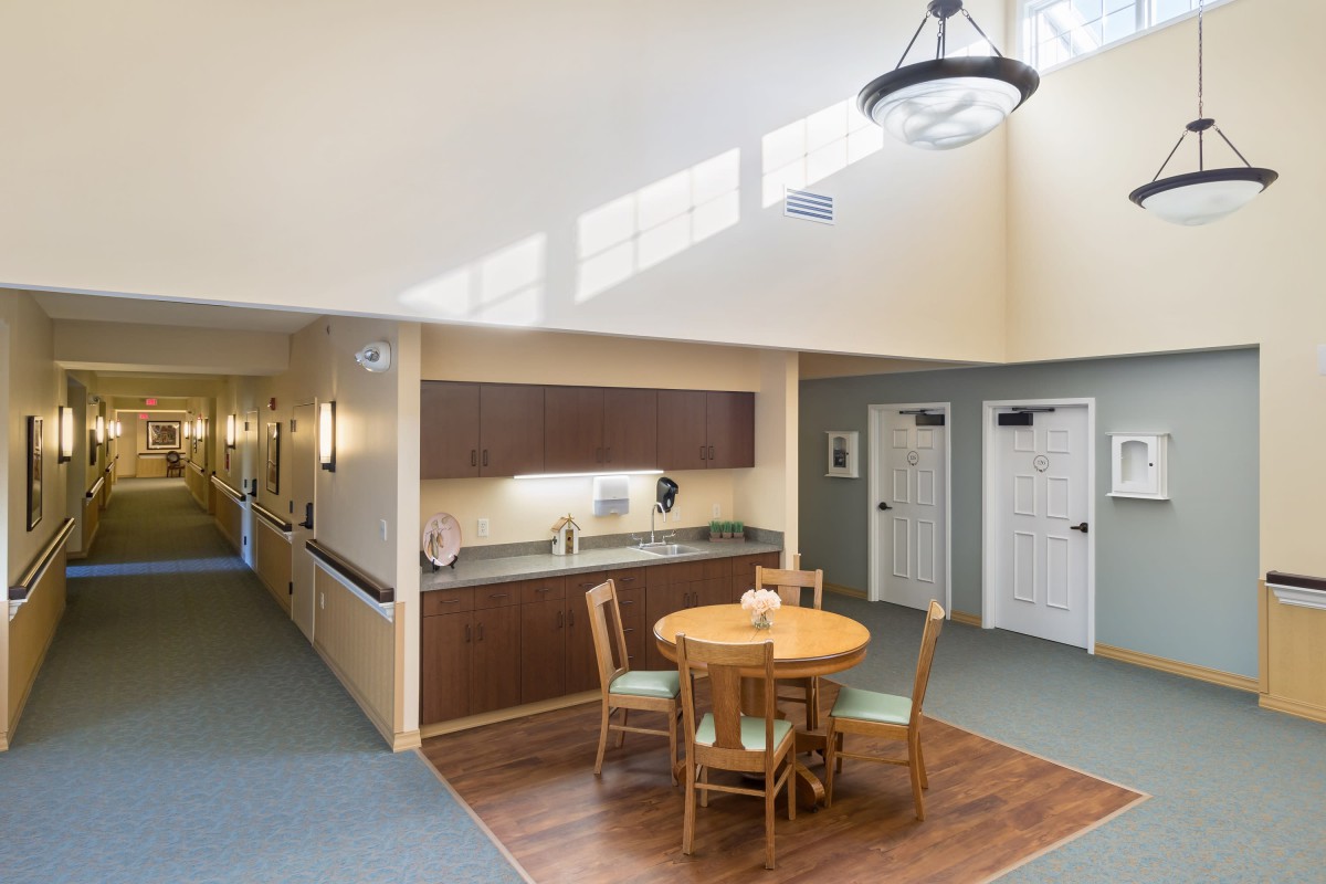 A minimalist interior featuring a dining area with a round table and four chairs, adjacent to a kitchenette and hallway.