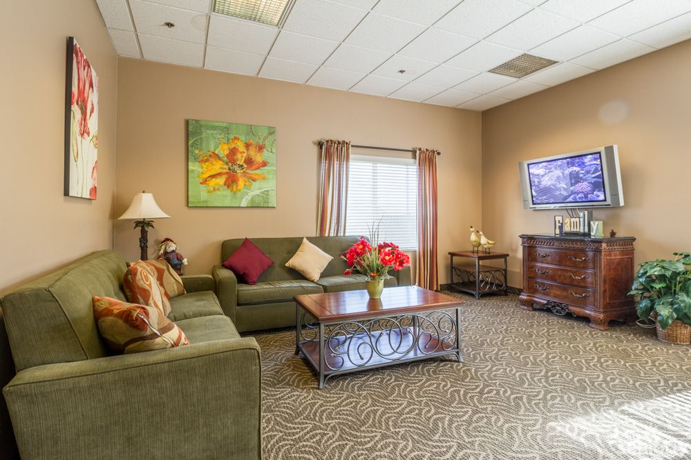 A cozy living room with a green sofa, decorative pillows, and a coffee table adorned with flowers. The room features a television, a colorful painting on the wall, and soft lighting provided by a floor lamp.