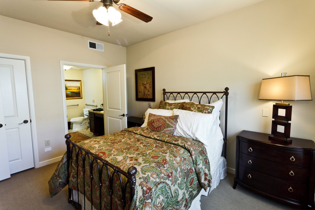 A warmly decorated bedroom featuring a double bed with patterned bedding, a bedside lamp, and an open doorway leading to another room.