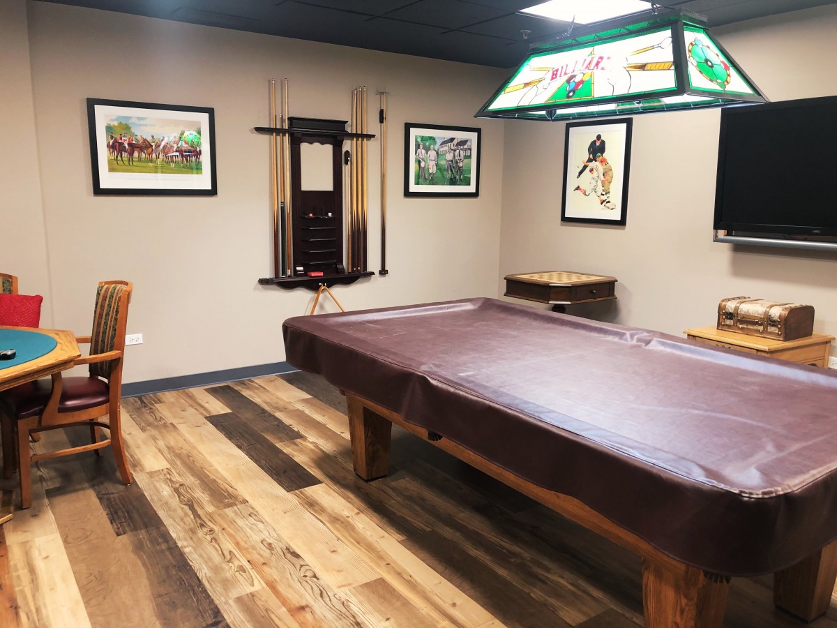 A cozy game room featuring a pool table with a cover, surrounded by framed artwork and warm wood flooring.