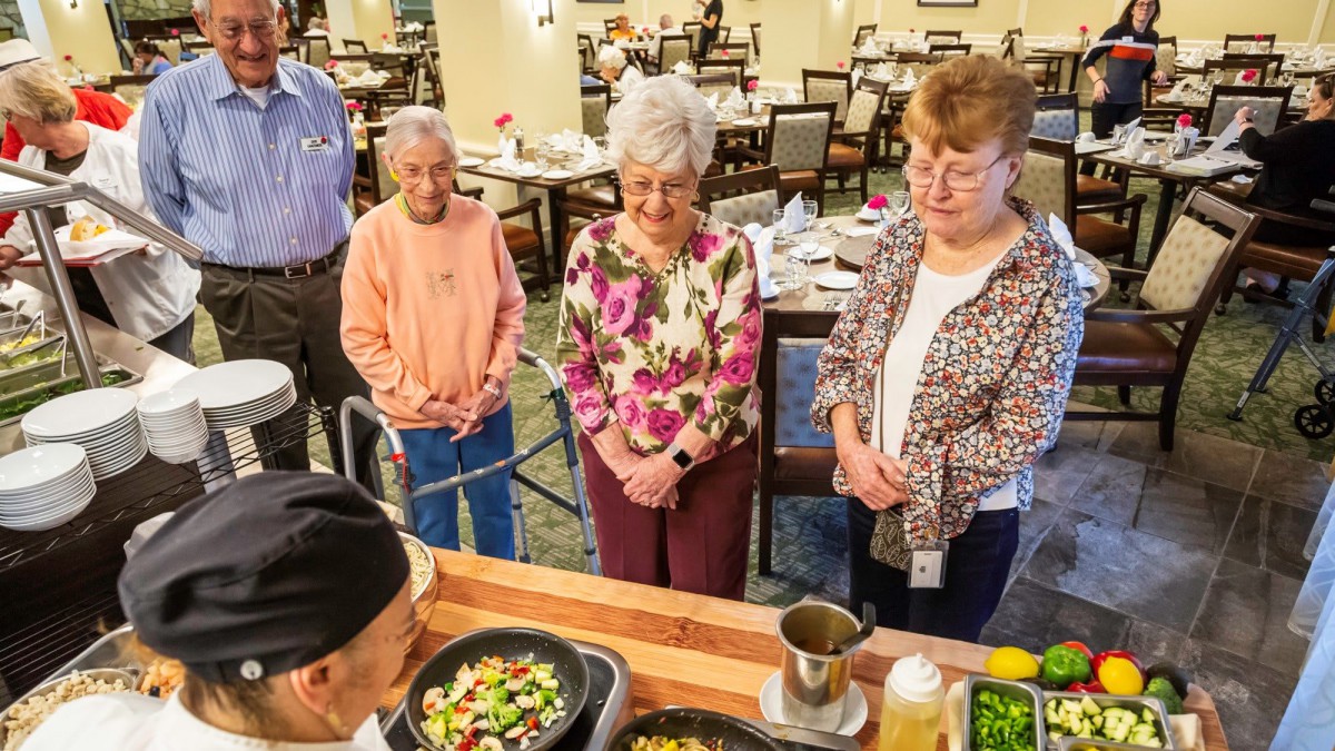 A group of elderly individuals watches a chef preparing food in a communal kitchen setting.