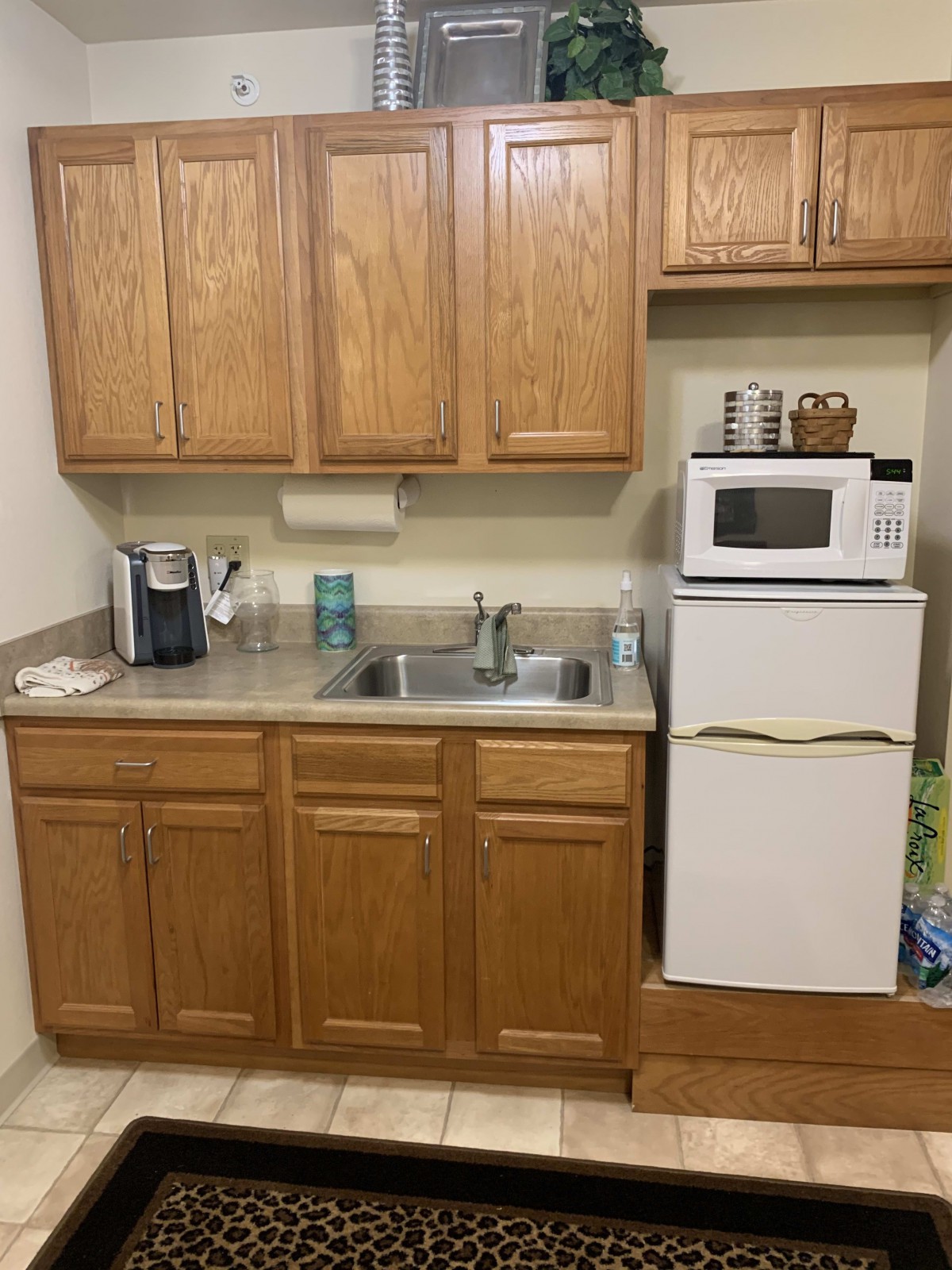 A compact kitchen area with wooden cabinets, a sink, a microwave on top of a mini fridge, and a coffee maker.