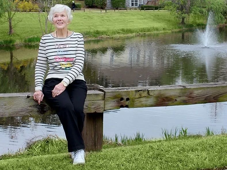 An elderly woman sitting on a bench by a pond in a park.