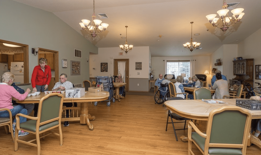 A nursing home dining area with tables, chairs, and residents seated during meal time.