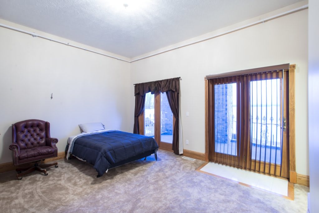A spacious bedroom with large windows, featuring a bed with dark bedding, carpet flooring, and wooden furnishings.