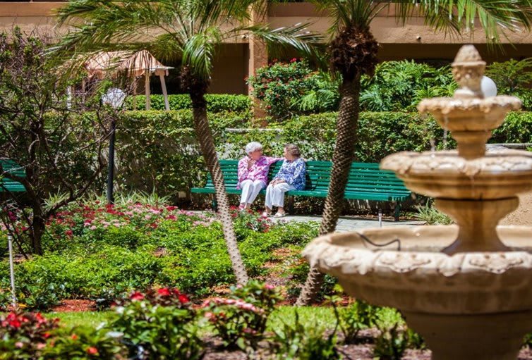 Two seniors sitting on a bench in a lush garden near a fountain.