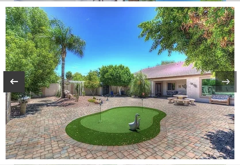 A spacious backyard with a putting green, surrounded by trees and outdoor seating on a paved patio.