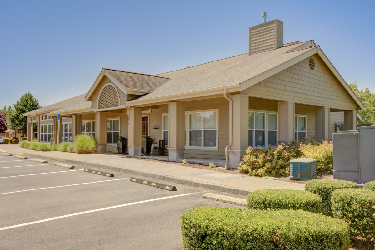 A suburban residential building with a parking lot in front, surrounded by neatly trimmed bushes under a clear sky.