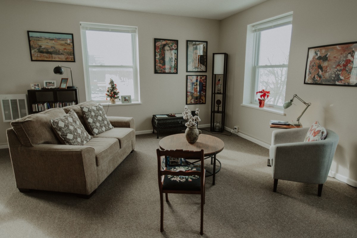 A cozy living room with a sofa, coffee table, and wall art, illuminated by natural light from two windows.