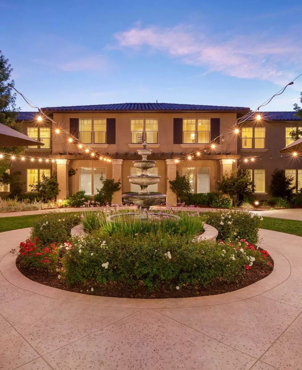 A beautifully lit courtyard features a central fountain surrounded by lush gardens at dusk.
