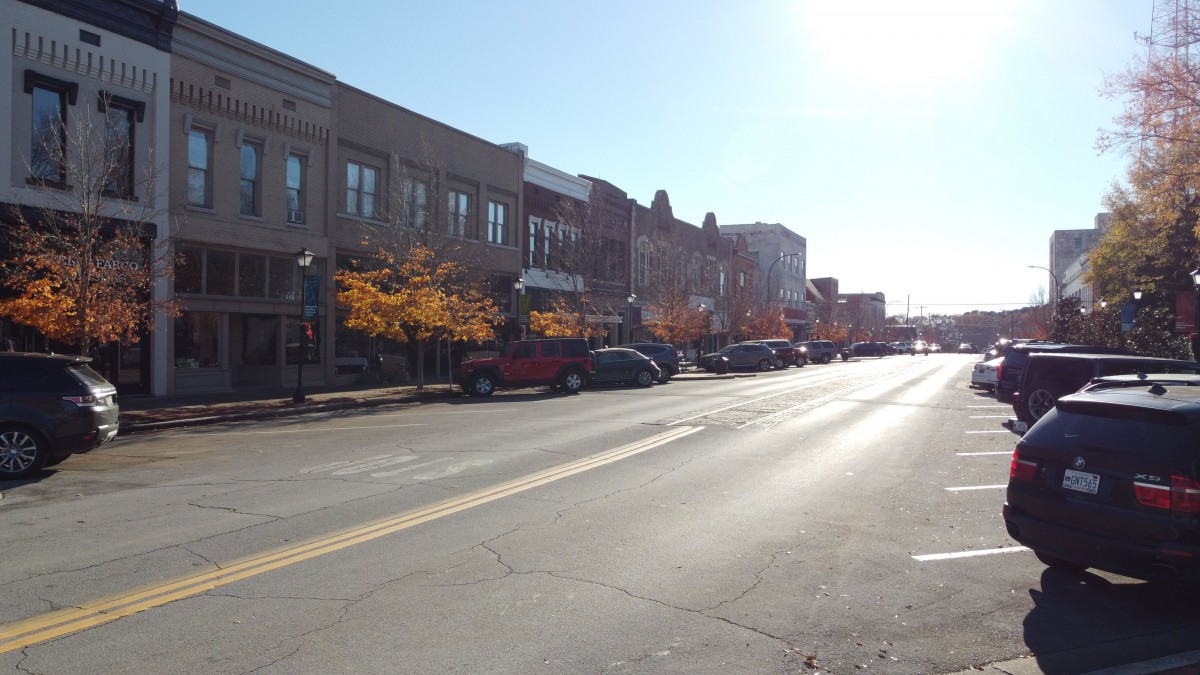 A street view lined with buildings, parked cars, and autumn trees under a clear sky.