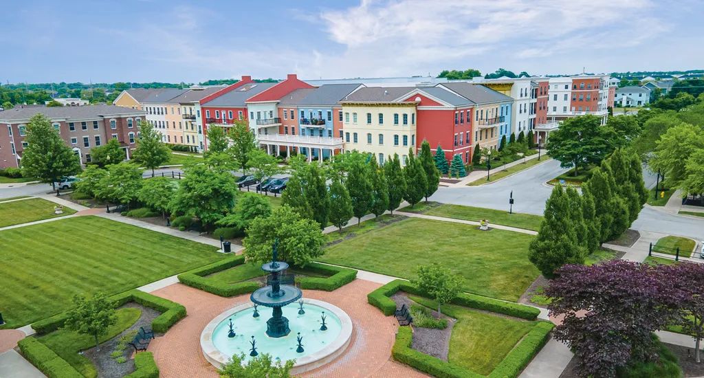 A serene park with a central fountain, surrounded by colorful buildings and lush greenery.