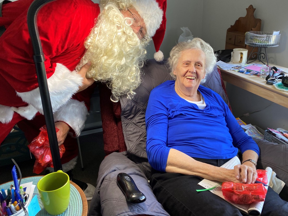 An elderly woman smiles while sitting next to a person dressed as Santa Claus.